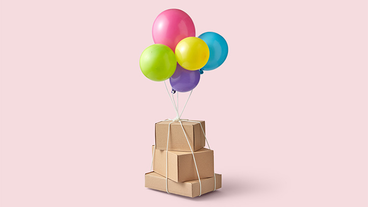 cardboard boxes tied to five colorful balloons in green, yellow, blue, pink and purple against a pink background